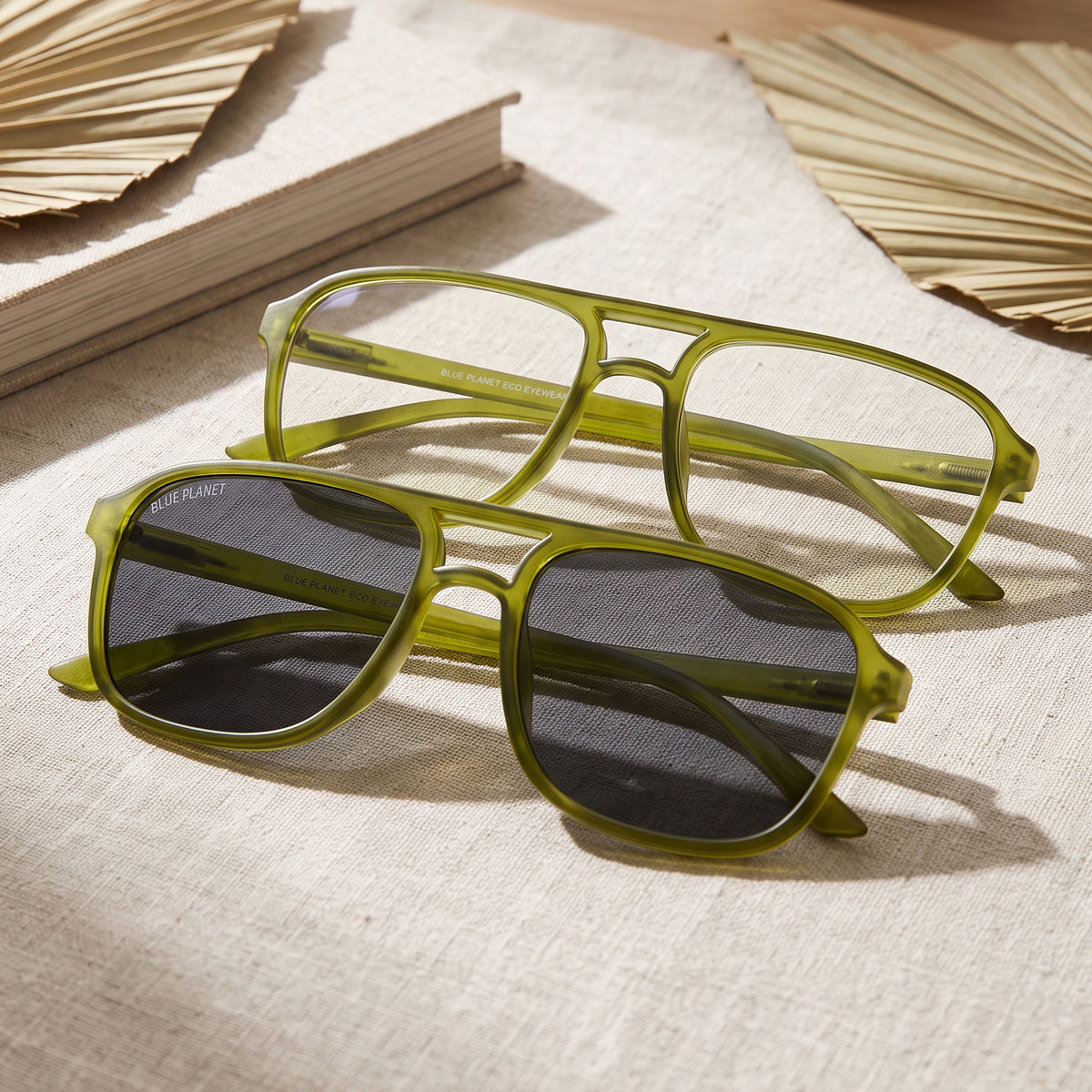 A pair of square aviator-style sunglasses with translucent olive green frames and dark gray lenses, displayed on a textured beige fabric alongside a book and dried palm leaves.