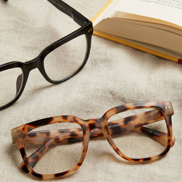 Two pairs of eyeglasses, one black and one tortoiseshell, on a textured surface with a book in the background.