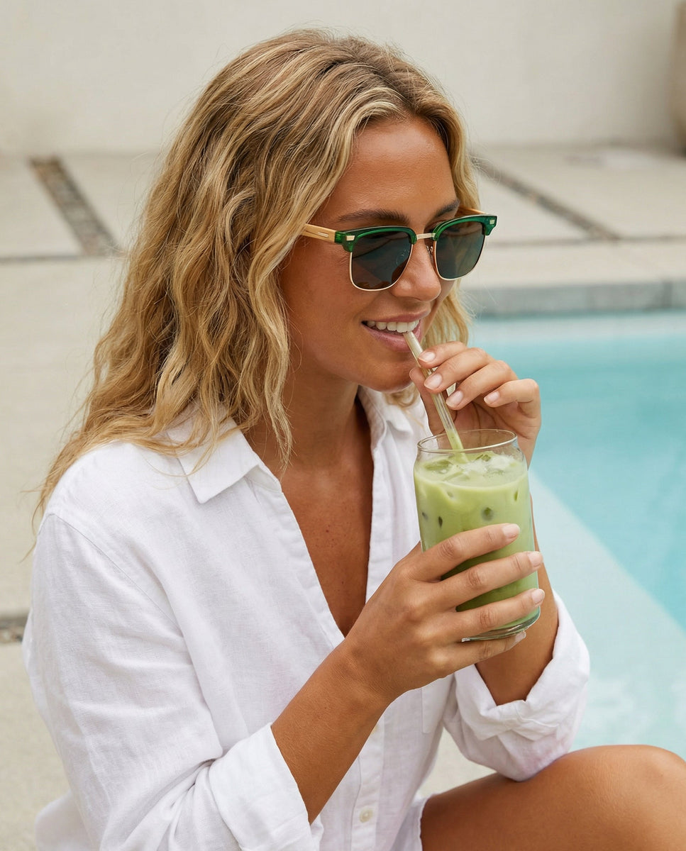 Woman in white shirt and sunglasses drinking a matcha by a pool.